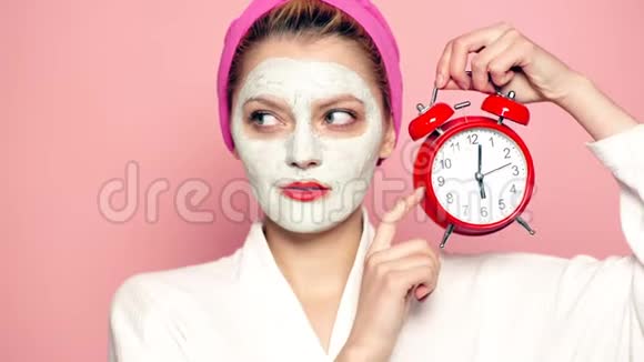 一个女孩脸上戴着面具头上戴着毛巾手里拿着一个闹钟背景是粉红色的面部护理视频的预览图
