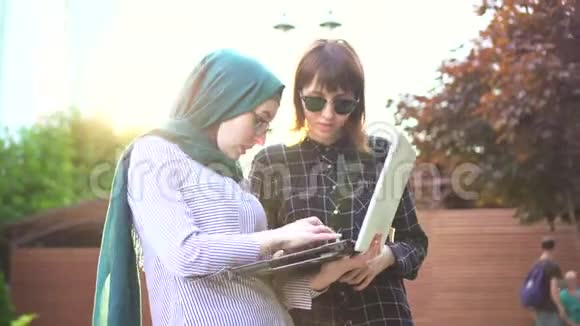 穆斯林女学生戴着头巾她的朋友带着笔记本电脑在公园里聊天阳光明媚视频的预览图