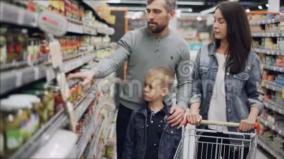 已婚夫妇带着可爱的小儿子在买食物他们从货架上拿着玻璃罐检查食材视频的预览图