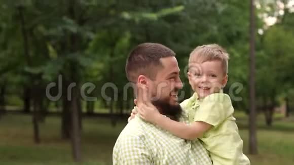 年轻的爸爸抱着他可爱的小儿子在公园的绿树背景视频的预览图