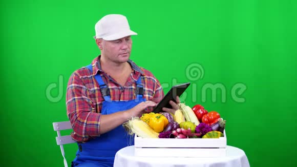 穿着格子衬衫和蓝色连衣裙的英俊的男农民手里拿着一块Chromakey绿色背景和盒子上的平板电脑视频的预览图