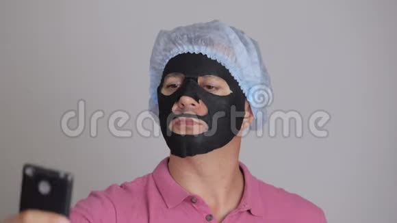 一个男人把一个恢复活力的黑布化妆品面膜敷在脸上用智能手机自拍视频的预览图