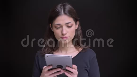 集中的白种人长发黑发女孩的肖像在黑色背景下专注地观看平板电脑视频的预览图