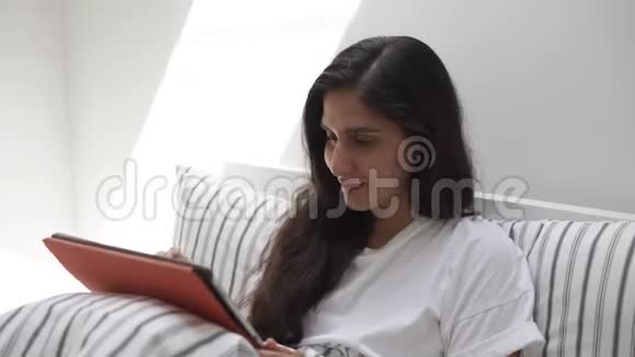 年轻的黑发女郎高兴地看着躺在床上的平板电脑屏幕视频的预览图