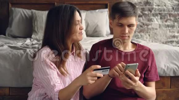 英俊的年轻人正在用智能手机购买互联网而他迷人的妻子则坐在他身边视频的预览图