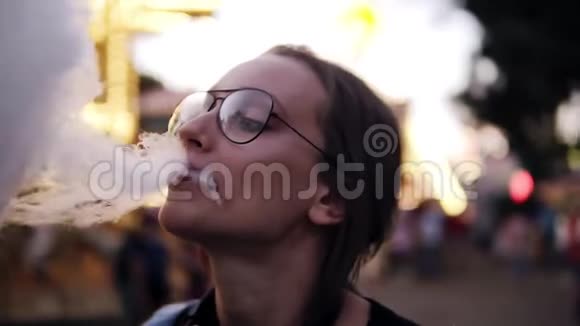 美丽的女孩吃棉花糖在游乐园附近模糊的旋转木马戴着时髦的眼镜很开心视频的预览图