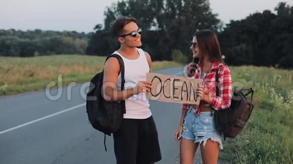 年轻的旅行夫妇相爱站在路上举着一个标志海洋快乐的微笑的朋友们正在注视视频的预览图