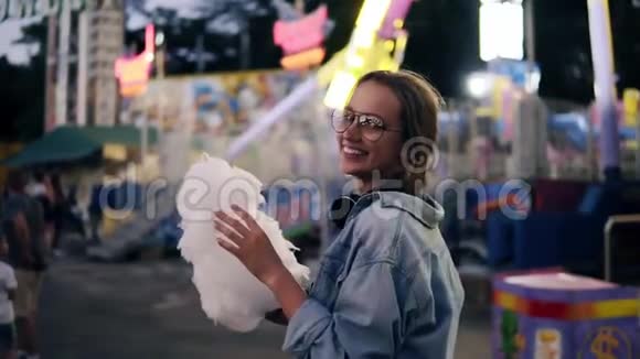 一个年轻的金发女孩在娱乐公园里散步吃着白色的棉花糖看着她视频的预览图
