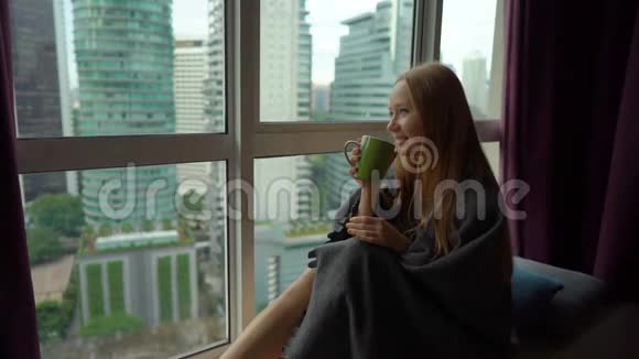 超级慢动作镜头一个年轻的女人喝茶或咖啡包裹在格子坐在全景窗口视频的预览图