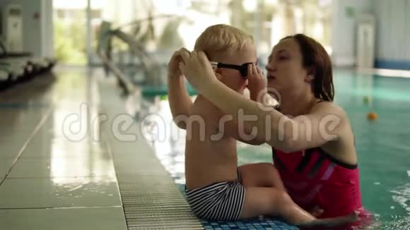 一个小游泳运动员准备游泳这个男孩坐在游泳池的边缘他妈妈戴上眼镜游泳视频的预览图