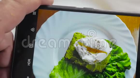 餐馆里的一个男人用手机摄像头拍摄食物荷包蛋三明治和鳄梨视频的预览图