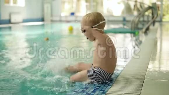 一个金发的小男孩坐在水池边脚在水里弹用脚泼水侧面视频的预览图