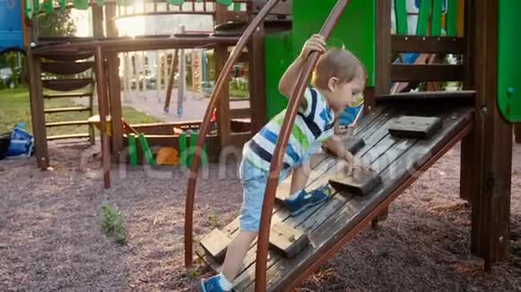 在公园的操场上一个蹒跚学步的小男孩挣扎着爬上木梯视频的预览图
