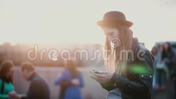美丽的年轻欧洲妇女戴着时尚的帽子使用智能手机应用程序在模糊繁忙的日落街道上慢动作视频的预览图