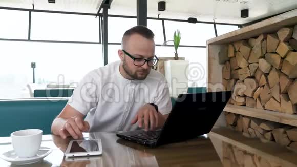 一个留胡子的年轻人戴着眼镜在笔记本电脑和平板电脑上工作休息一下他喝咖啡快节奏概念视频的预览图