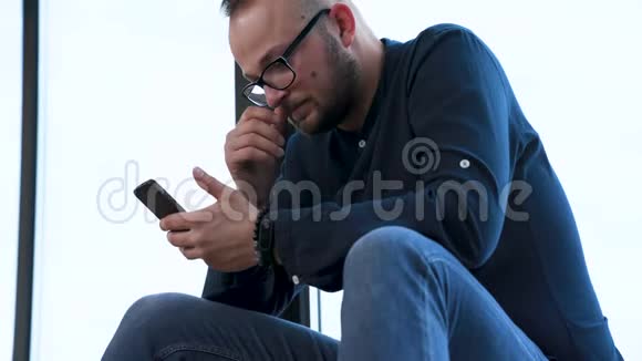 一个年轻的悲伤男人坐在全景窗口附近翻过手机上的屏幕视频的预览图