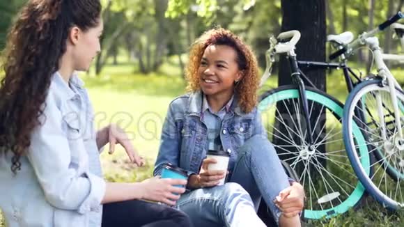 两个女骑自行车的人正在休息坐在公园的草坪上喝咖啡骑自行车后聊天积极主动视频的预览图