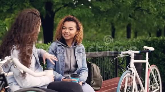 年轻的女游客正坐在公园的长椅上端着带着自行车的外卖咖啡站着聊天视频的预览图