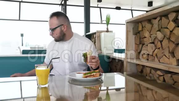 一个长着胡子的年轻人戴着眼镜吃三明治喝橙汁人在lucnh接电话视频的预览图