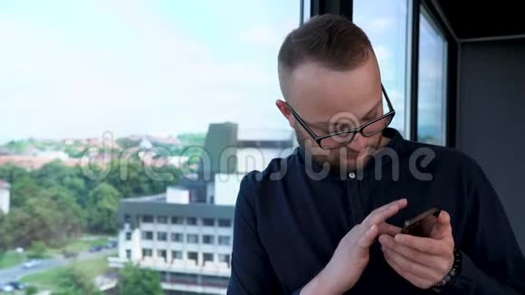 一个戴着眼镜的留胡子的年轻人呆在窗户附近他看着他的手机和打字信息视频的预览图