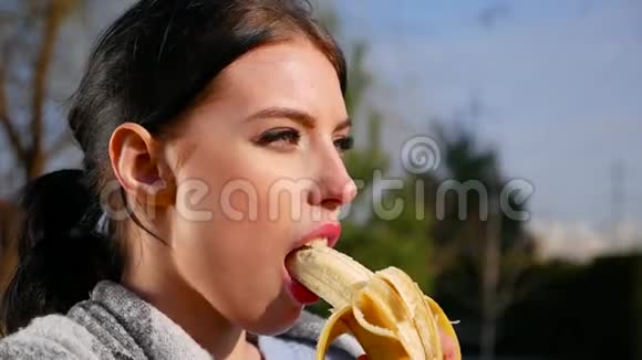 在阳光明媚的日子里有着美丽的眼睛和红唇的迷人年轻黑发女人吃香蕉视频的预览图