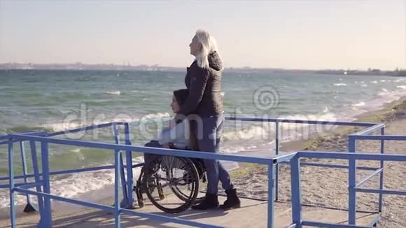 家庭休闲轮椅上的年轻残疾妇女和她的母亲一起在斜坡上的海边散步视频的预览图