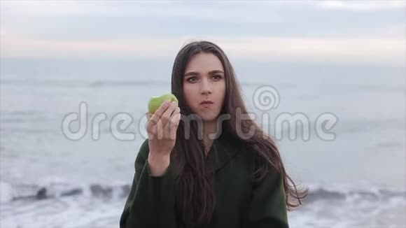 慢动作日出时穿着深绿色外套在海边吃苹果的漂亮女孩特写视频的预览图