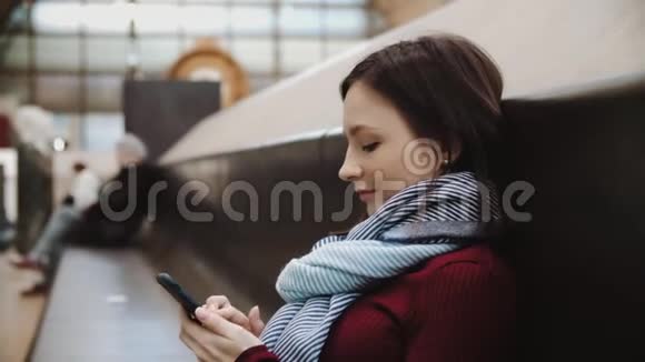 坐在火车站大理石长凳上的兴奋的女人在智能手机上发短信视频的预览图
