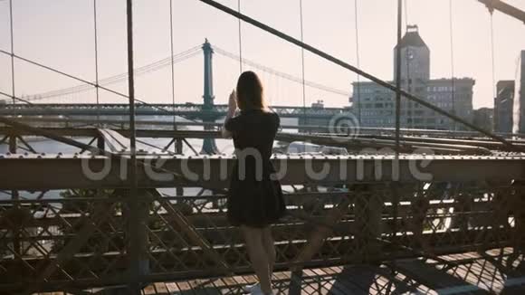 美丽的高加索女人沿着纽约布鲁克林大桥走停下来拍智能手机照片的风景4K视频的预览图
