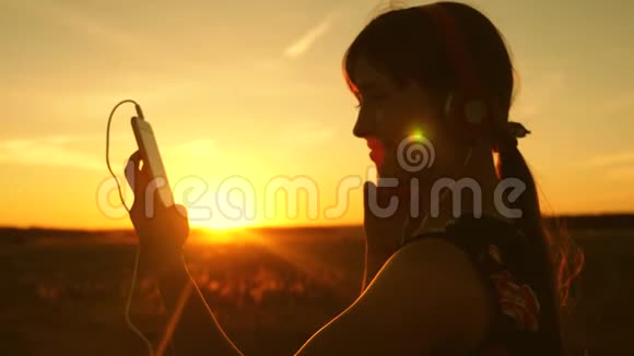 快乐的女孩在智能手机上听音乐在美丽的春天日落的光线下慢动作年轻女孩视频的预览图