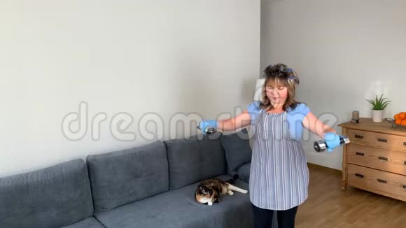 肥胖的中年妇女一个穿着围裙的家庭主妇在家里用哑铃做体育锻炼运动的概念体重视频的预览图