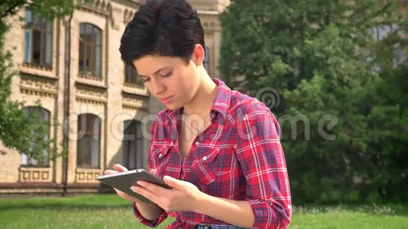 美丽的年轻女子留着黑色短发在平板电脑上打字对着镜头微笑坐在大学附近的公园里视频的预览图