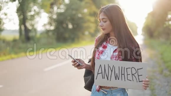 年轻漂亮的女人搭车站在路上拿着任何地方的标志用智能手机少女视频的预览图