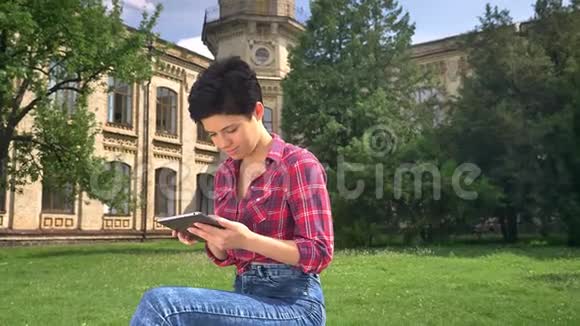 美丽快乐的女人留着黑色短发在平板电脑上打字在摄像机前微笑坐在大学附近的公园里视频的预览图