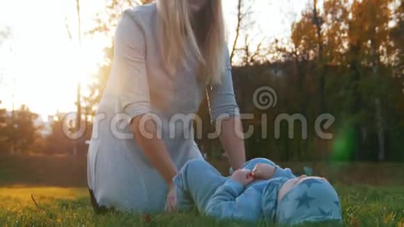 年轻的母亲带着她的小宝宝在日落时在秋天的公园里玩耍婴儿躺在草地上视频的预览图