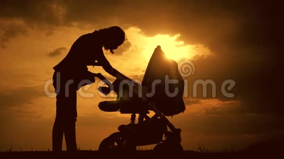 年轻母亲的剪影带着婴儿车在日落时享受母性年轻女子看着孩子视频的预览图