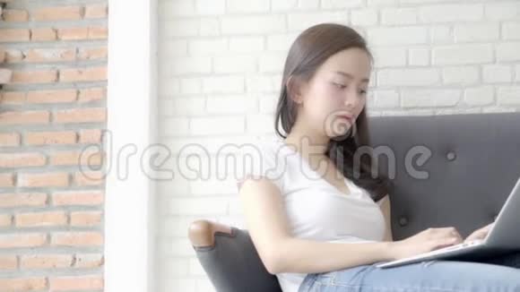 美丽的亚洲女人坐在沙发上在卧室里用笔记本电脑和智能手机聊天休闲放松视频的预览图