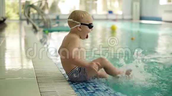 一个金发小男孩坐在游泳池的边缘穿着泳裤和眼镜用脚喷水视频的预览图