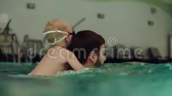 一位年轻的父亲背上有一个小儿子在水面上划船室内游泳池相机半在水里快乐的孩子视频的预览图