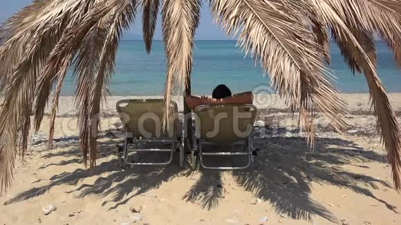 男人躺在日光浴床上的棕榈树遮阳伞下欣赏蔚蓝的大海在异国情调的海滩上放松度假视频的预览图