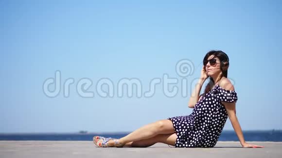 美丽的黑发女孩戴着太阳镜和日光浴带着大耳机在海滩上听智能手机上的音乐视频的预览图