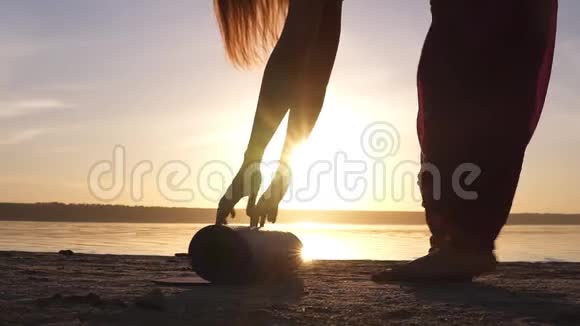 靠近一个瑜伽女子展开瑜伽垫在靠近水的沙子上阳光明媚清晨黄昏视频的预览图