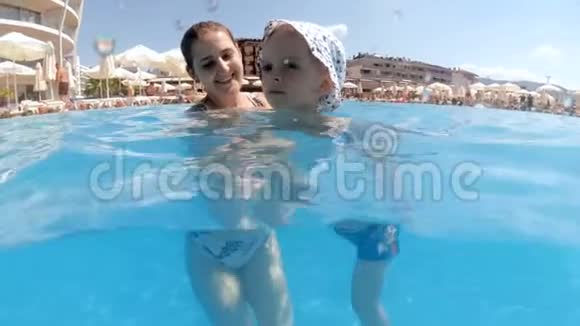 慢动作水下视频微笑的年轻女子与蹒跚学步的男孩游泳在酒店度假胜地的室外游泳池热视频的预览图