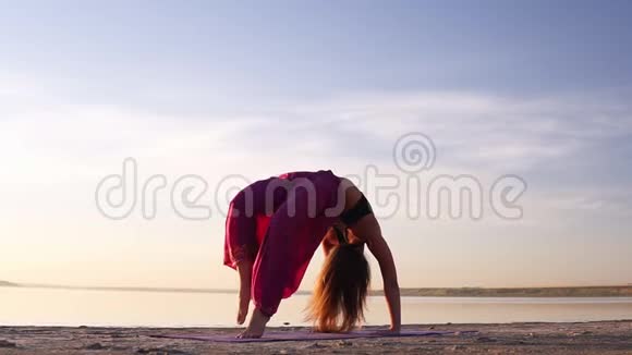早上一位穿着粉红色瑜伽裤的年轻女子在海边或湖边的沙滩上练习近距离观看姿势视频的预览图