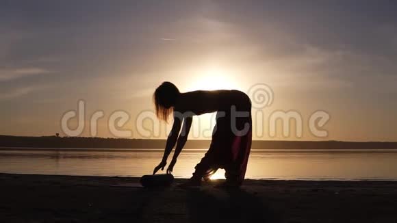 一个身材匀称的女人在水边的剪影展开瑜伽垫准备做瑜伽或普拉提日落海滩视频的预览图