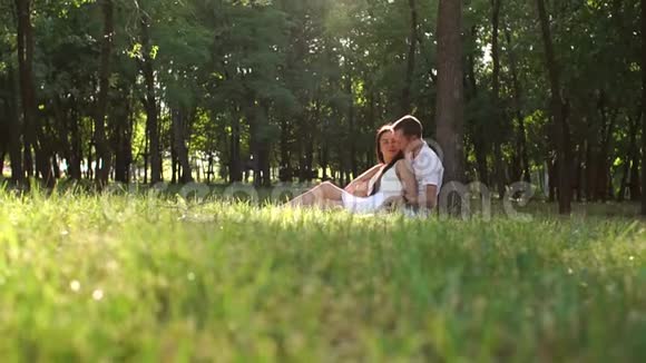 年轻的青少年恋人正坐在草地上靠着公园里的一棵树视频的预览图