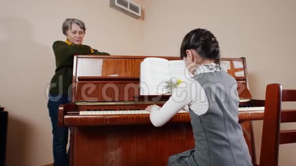 音乐课女孩弹钢琴年长的老师站在钢琴附近从女孩的背后看游戏的画面视频的预览图
