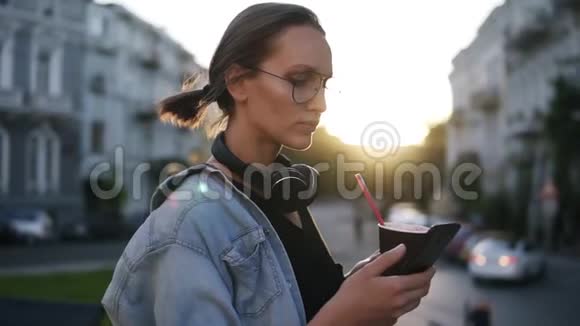 漂亮的年轻女孩穿着蓝色衬衫脖子上戴着耳机站在城市街道上滚动着她的手机视频的预览图