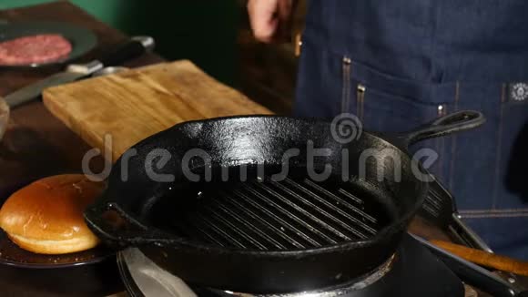 身着蓝色围裙的男厨师站在餐桌旁配有煎锅烤架和汉堡配料视频的预览图