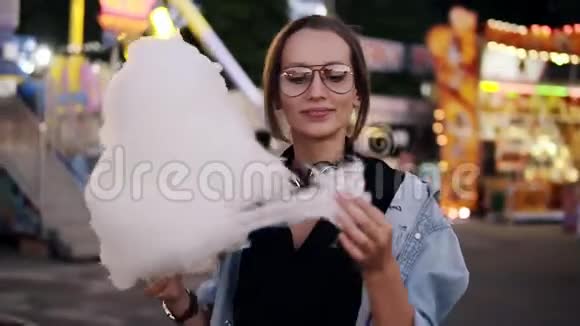 一个迷人的女孩走在娱乐公园吃白色棉花糖穿休闲装的女孩戴着耳机视频的预览图